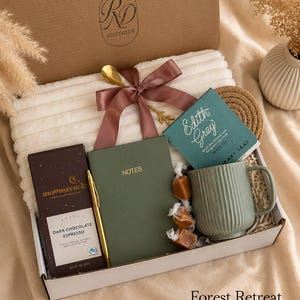 Peut inclure: Un coffret cadeau contenant une couverture blanche c&ocirc;tel&eacute;e avec un ruban mauve, une tablette de chocolat noir espresso Moonstruck, un carnet vert &eacute;tiquet&eacute; NOTES, un stylo dor&eacute;, une bo&icirc;te de th&eacute; Edith Grey, une tasse vert sauge et trois caramels.
