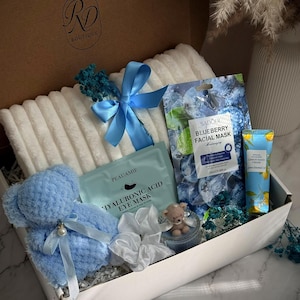 May include: A gift box labeled "RD Boutique" filled with a blue teddy bear, a white towel, face masks, and a tube of hand cream. The box is decorated with blue ribbons and dried flowers. The text "Blue Serenity" is at the bottom.