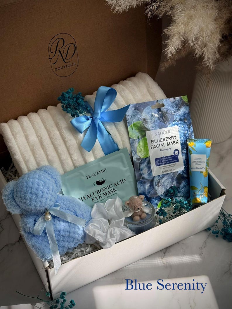 May include: A gift box labeled "RD Boutique" filled with a blue teddy bear, a white towel, face masks, and a tube of hand cream. The box is decorated with blue ribbons and dried flowers. The text "Blue Serenity" is at the bottom.
