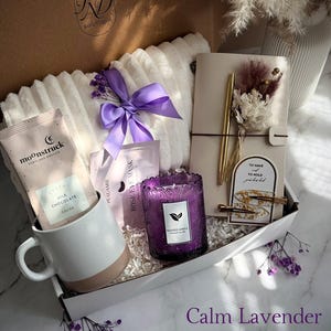 May include: A gift box with a white and beige mug, a purple candle, a notebook, and a white blanket. The box also contains cocoa packets, eye masks, and a purple bow. The text "Calm Lavender" is at the bottom.