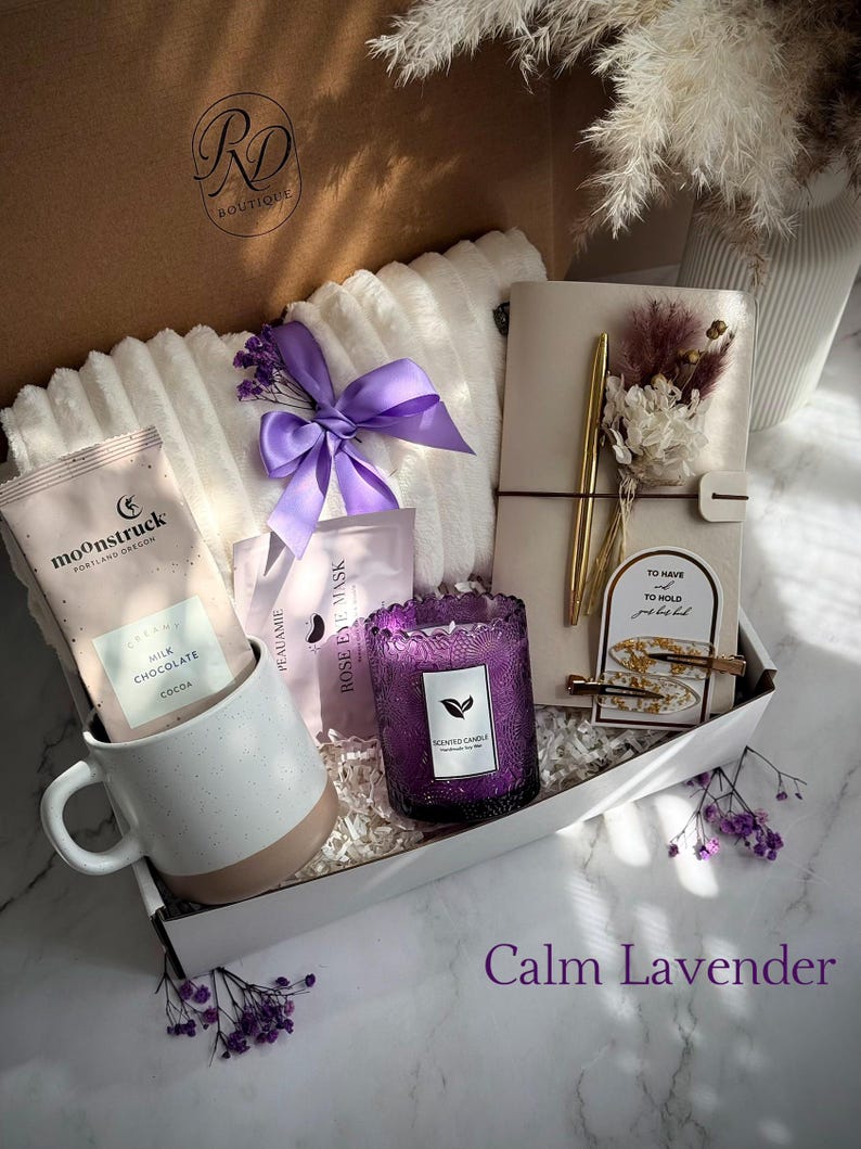 May include: A gift box with a white and beige mug, a purple candle, a notebook, and a white blanket. The box also contains cocoa packets, eye masks, and a purple bow. The text "Calm Lavender" is at the bottom.