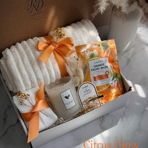 May include: A gift box with white towels tied with orange ribbons, a scented candle, orange facial mask, and decorative accents. The box is labeled "RD Boutique" and the text "Citrus Glow" is visible.