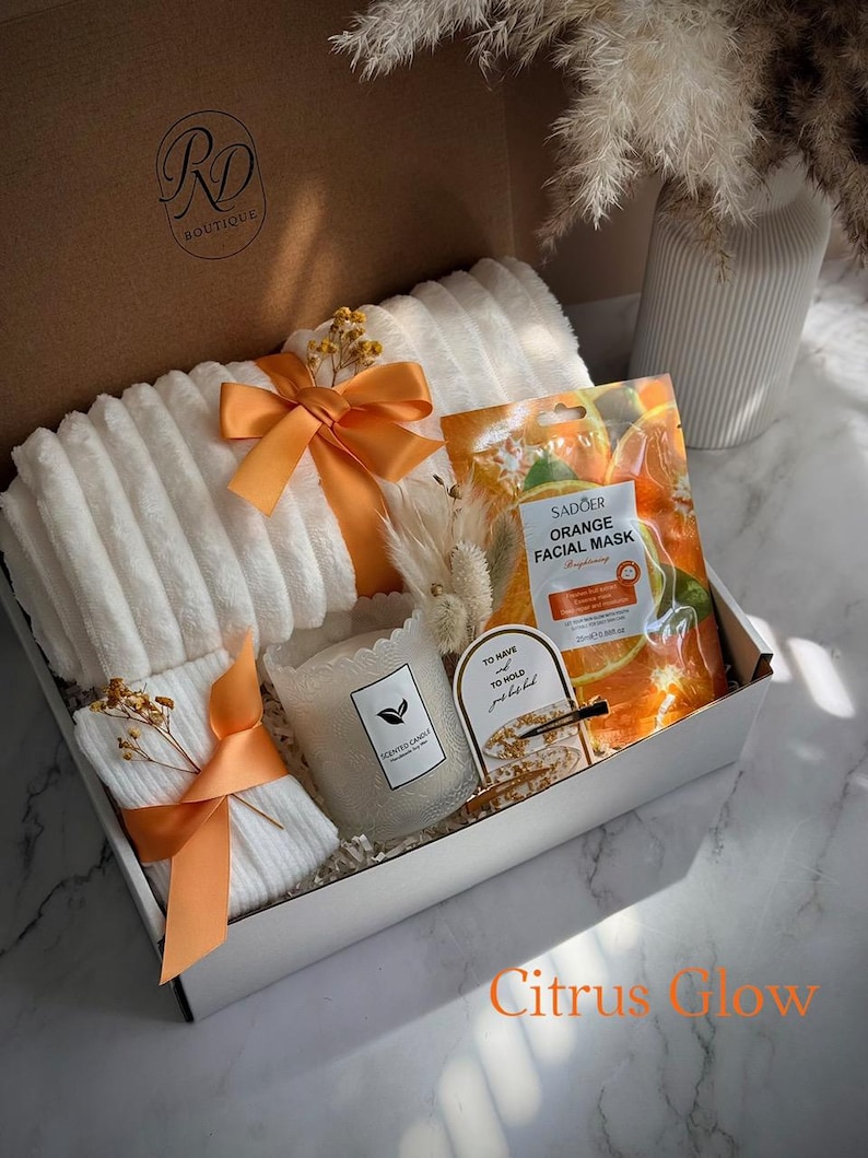 May include: A gift box with white towels tied with orange ribbons, a scented candle, orange facial mask, and decorative accents. The box is labeled "RD Boutique" and the text "Citrus Glow" is visible.
