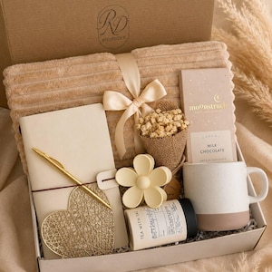 Peut inclure: Un coffret cadeau contenant une couverture c&ocirc;tel&eacute;e beige avec un ruban en satin, un carnet cr&egrave;me avec un stylo dor&eacute;, un ornement en fil dor&eacute; en forme de c&oelig;ur, un petit bouquet de fleurs s&eacute;ch&eacute;es, une pince &agrave; cheveux fleur, une bo&icirc;te de th&eacute; et du chocolat.