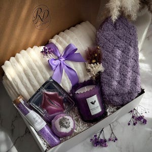 May include: A gift box filled with lavender-themed self-care items. Includes a white plush towel, a purple bath mitt, a candle, bath salts, a bath bomb, and a lip mask. The box is tied with a purple ribbon and has the text "Lavender Bliss".