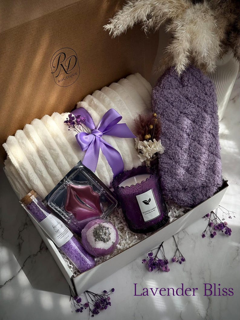 May include: A gift box filled with lavender-themed self-care items. Includes a white plush towel, a purple bath mitt, a candle, bath salts, a bath bomb, and a lip mask. The box is tied with a purple ribbon and has the text "Lavender Bliss".