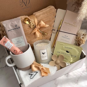 Peut inclure: Un coffret cadeau de bien-être contenant une tasse, une bougie, du chocolat, un masque pour les yeux, une crème pour les mains et une couverture douce. La boîte est beige et blanche avec un stylo et une cuillère dorés. Le texte "Luxe Calm Collection" est visible.
