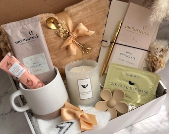 May include: A gift box filled with self-care items. Includes a mug, candle, chocolate, eye mask, hand cream, and a soft blanket. The box is beige and white with a gold pen and spoon. The text "Luxe Calm Collection" is visible.