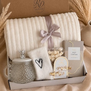 Peut inclure: Un coffret cadeau contenant un plaid c&ocirc;tel&eacute; cr&egrave;me nou&eacute; d'un ruban gris, une tasse en verre avec un couvercle en forme d'ours, une chaussette duveteuse blanche avec un c&oelig;ur noir, des guimauves, du chocolat chaud et deux pinces &agrave; cheveux dor&eacute;es.
