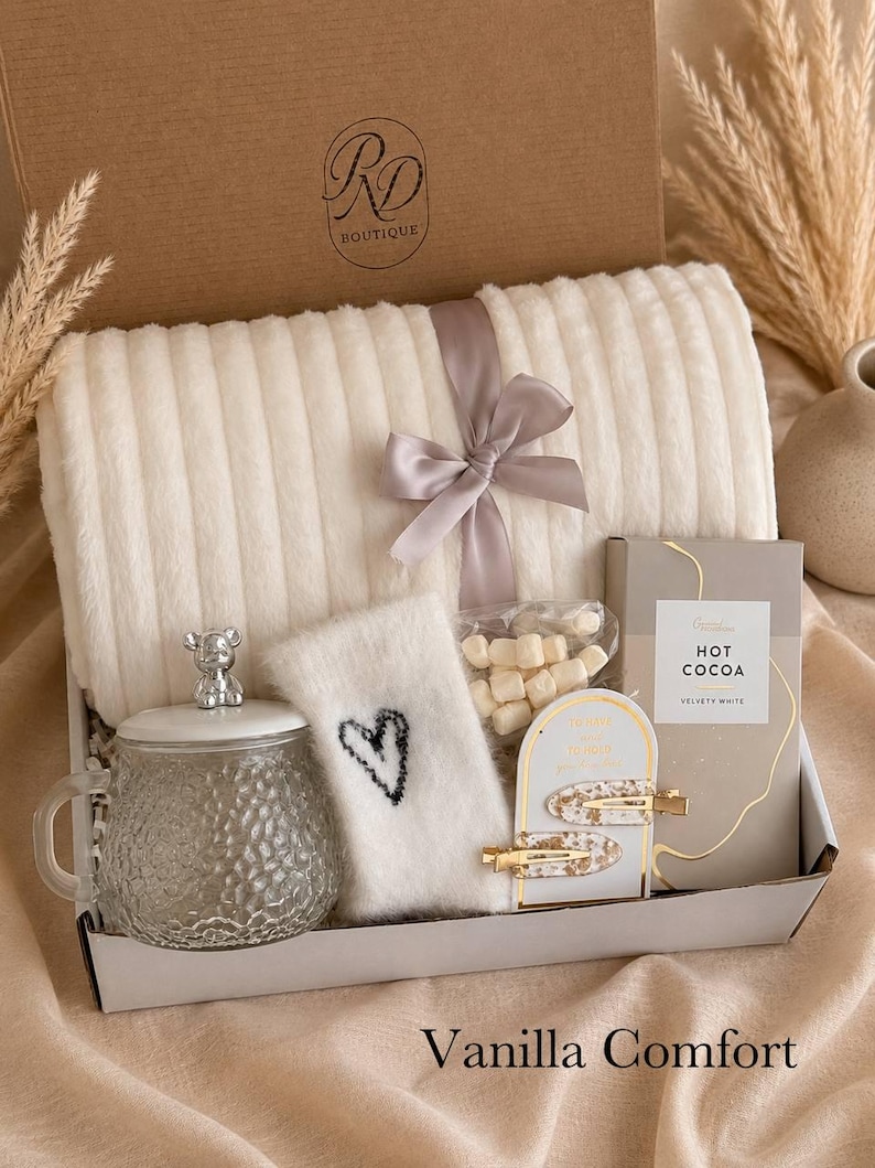 Peut inclure: Un coffret cadeau contenant un plaid c&ocirc;tel&eacute; cr&egrave;me nou&eacute; d'un ruban gris, une tasse en verre avec un couvercle en forme d'ours, une chaussette duveteuse blanche avec un c&oelig;ur noir, des guimauves, du chocolat chaud et deux pinces &agrave; cheveux dor&eacute;es.