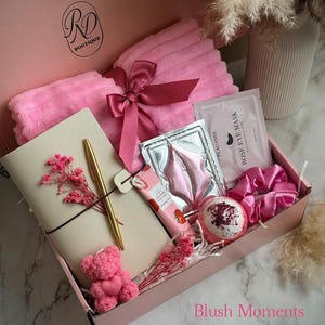May include: A pink gift box filled with self-care items. Includes a pink towel tied with a ribbon, a notebook, a gold pen, a teddy bear, eye masks, a bath bomb, a scrunchie, and a strawberry-scented lotion. The box has the text "Blush Moments".