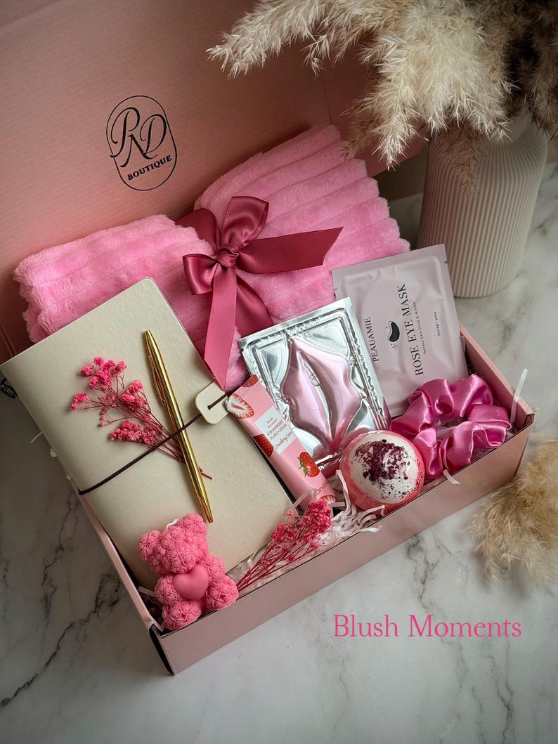 May include: A pink gift box filled with self-care items. Includes a pink towel tied with a ribbon, a notebook, a gold pen, a teddy bear, eye masks, a bath bomb, a scrunchie, and a strawberry-scented lotion. The box has the text "Blush Moments".