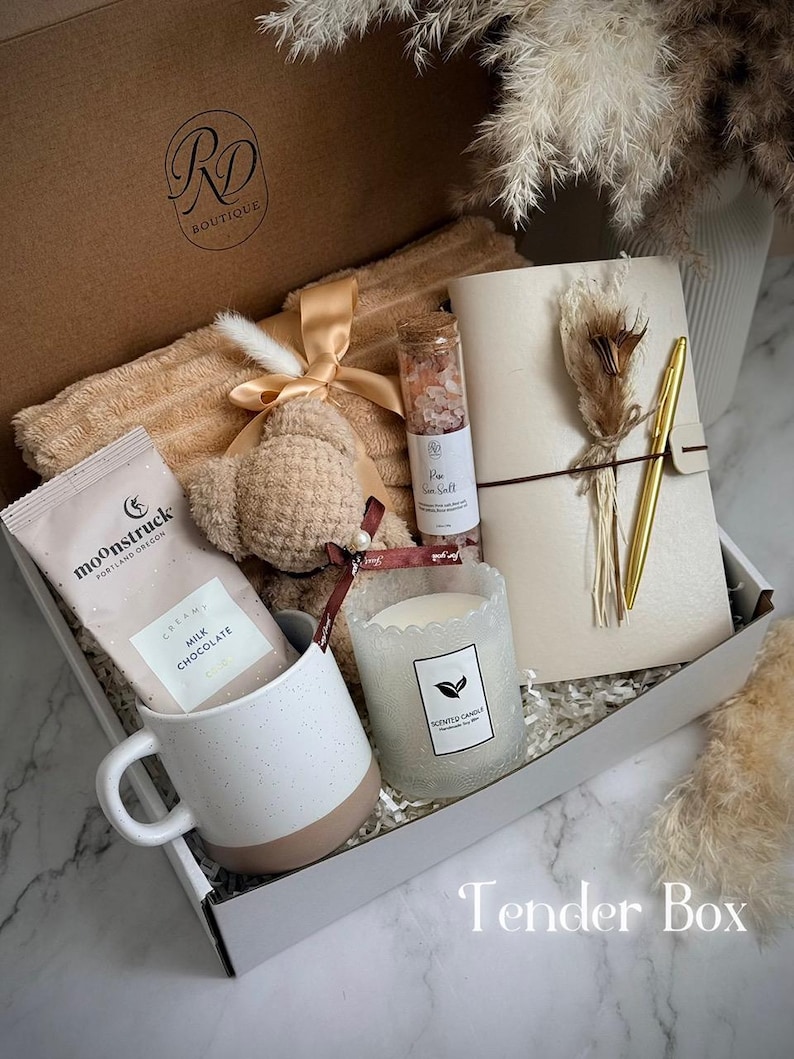 May include: A gift box with a variety of items, including a beige blanket, a teddy bear, a mug, a candle, a notebook, a pen, and a chocolate bar. The box is labeled "Tender Box."