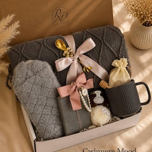 Peut inclure: Un coffret cadeau contenant une couverture tricot&eacute;e gris fonc&eacute; avec un ruban rose p&acirc;le, une pochette matelass&eacute;e grise, un carnet gris avec un n&oelig;ud rose et une breloque en perles, un pot de miel, une cuill&egrave;re en bois, un mug noir et une fleur.
