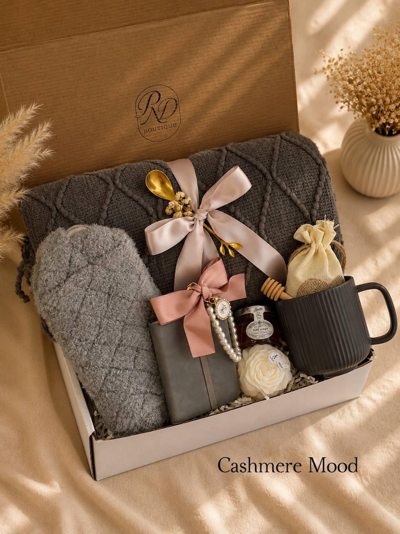 Peut inclure: Un coffret cadeau contenant une couverture tricot&eacute;e gris fonc&eacute; avec un ruban rose p&acirc;le, une pochette matelass&eacute;e grise, un carnet gris avec un n&oelig;ud rose et une breloque en perles, un pot de miel, une cuill&egrave;re en bois, un mug noir et une fleur.