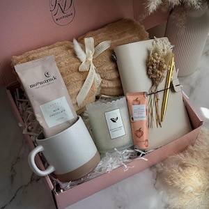 May include: A pink gift box filled with a beige blanket, a white and brown mug, a packet of cocoa, a candle, a notebook, a pen, and a tube of hand cream. The box has the text "RD Boutique" and "Soft Peach".