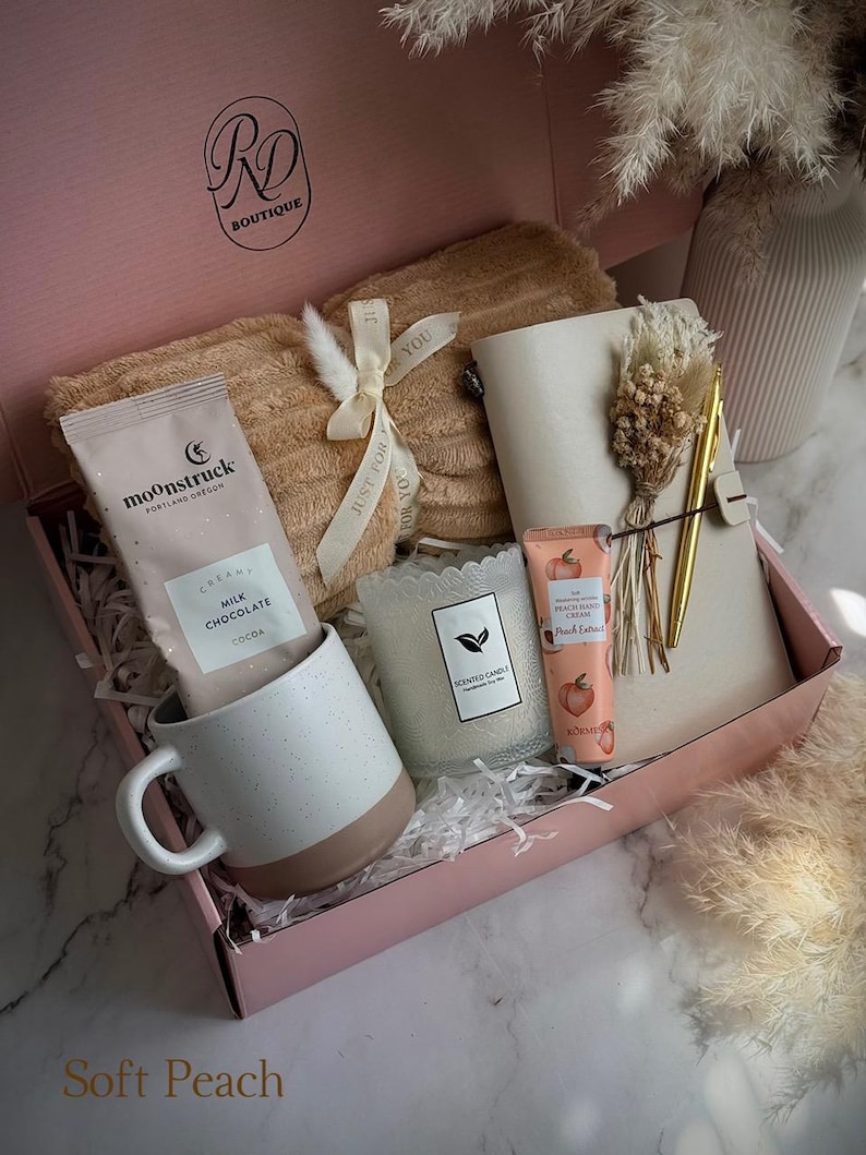 May include: A pink gift box filled with a beige blanket, a white and brown mug, a packet of cocoa, a candle, a notebook, a pen, and a tube of hand cream. The box has the text "RD Boutique" and "Soft Peach".