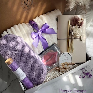May include: A gift box filled with self-care items. Includes a fluffy white pillow with a purple ribbon, a purple eye mask, a notebook with a gold pen, a glass tube of sea salt, and lip-shaped soap. The box is labeled "Purple Escape".