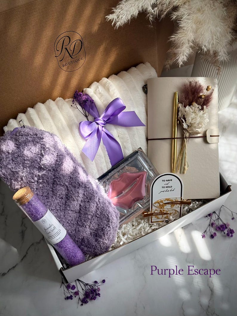 May include: A gift box filled with self-care items. Includes a fluffy white pillow with a purple ribbon, a purple eye mask, a notebook with a gold pen, a glass tube of sea salt, and lip-shaped soap. The box is labeled "Purple Escape".
