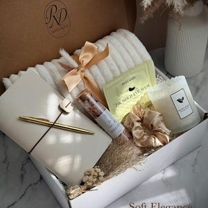 May include: A gift box filled with self-care items. Includes a white plush blanket, a journal with a gold pen, a candle, eye masks, bath salts, and a satin scrunchie. The box is tied with a peach ribbon. The text "Soft Elegance" is visible.