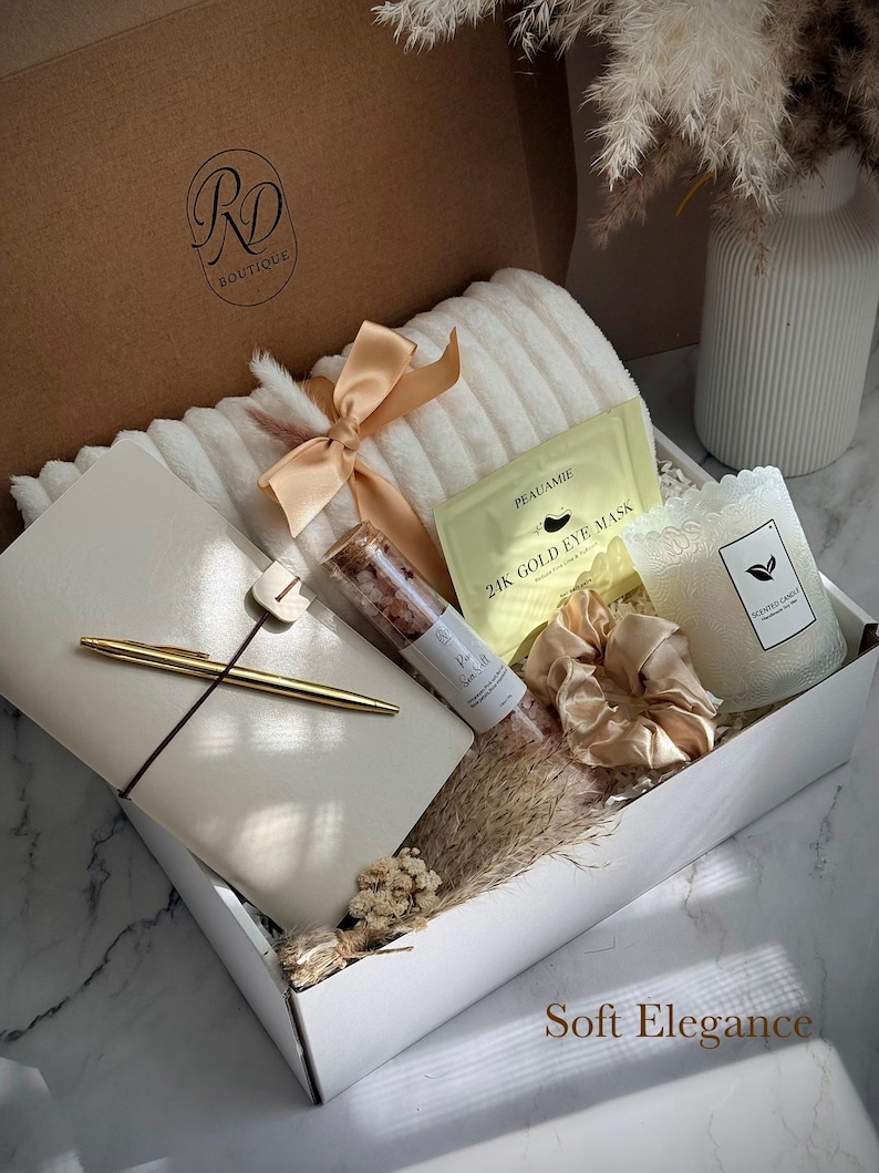 May include: A gift box filled with self-care items. Includes a white plush blanket, a journal with a gold pen, a candle, eye masks, bath salts, and a satin scrunchie. The box is tied with a peach ribbon. The text "Soft Elegance" is visible.