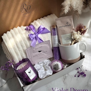 May include: A gift box filled with self-care items. Includes a white blanket with a purple bow, a purple candle, a mug, bath products, and a face mask. The box is labeled "Violet Dream".