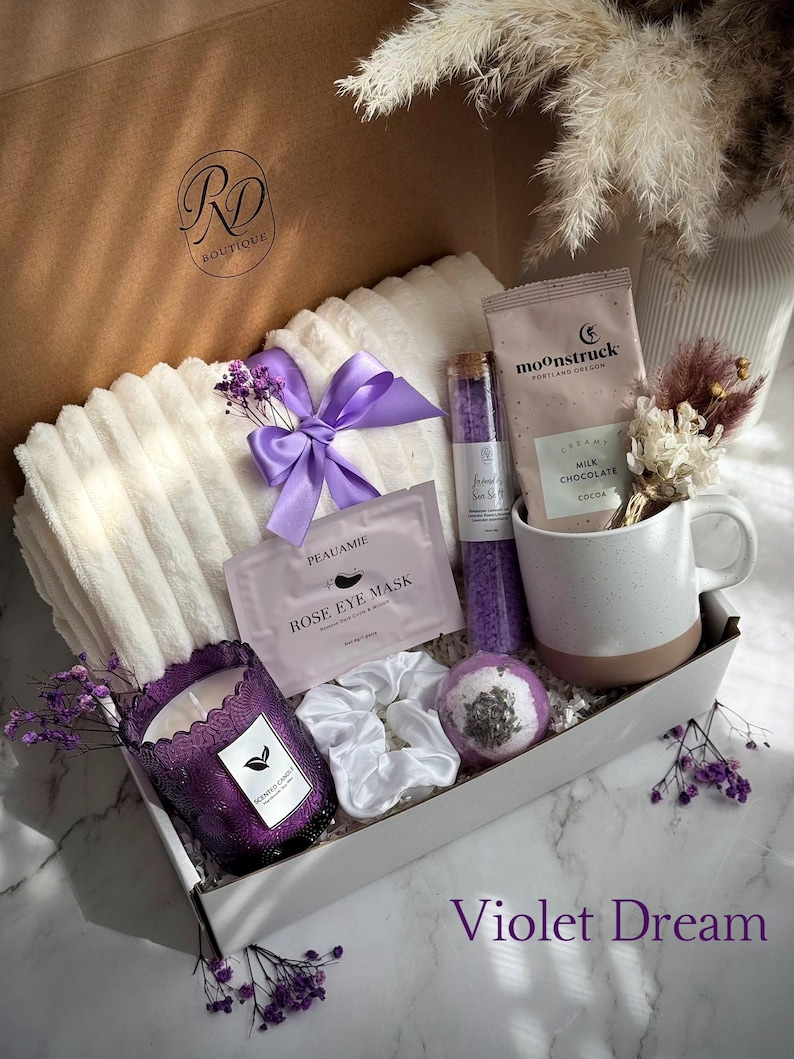 May include: A gift box filled with self-care items. Includes a white blanket with a purple bow, a purple candle, a mug, bath products, and a face mask. The box is labeled "Violet Dream".