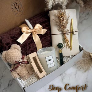 May include: A gift box with a brown teddy bear, a brown blanket with a gold bow, a journal with a gold pen, a candle, a hair clip, and a jade roller. The box has the text "Deep Comfort".