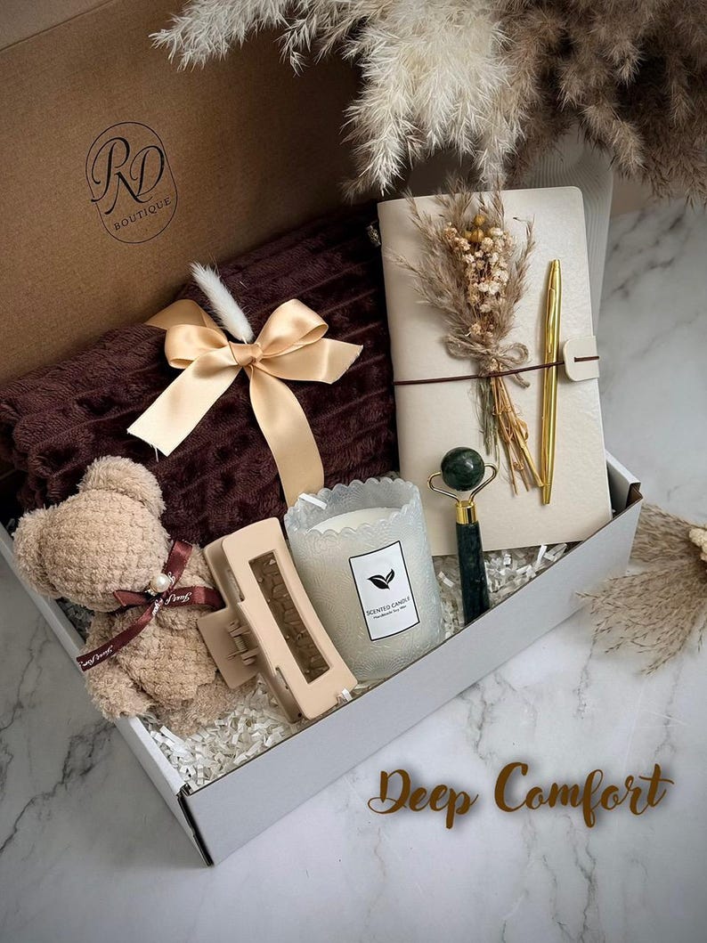 May include: A gift box with a brown teddy bear, a brown blanket with a gold bow, a journal with a gold pen, a candle, a hair clip, and a jade roller. The box has the text "Deep Comfort".