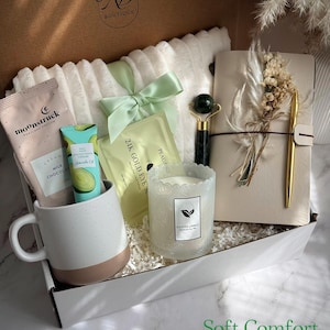 May include: A gift box filled with self-care items. Includes a white mug with a brown base, a face roller, a candle, a notebook with a pen, eye masks, and skincare products. The box is tied with a green ribbon and has the text "Soft Comfort".