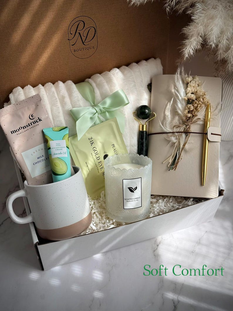 May include: A gift box filled with self-care items. Includes a white mug with a brown base, a face roller, a candle, a notebook with a pen, eye masks, and skincare products. The box is tied with a green ribbon and has the text "Soft Comfort".