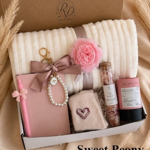 Peut inclure: Un coffret cadeau contenant un plaid c&ocirc;tel&eacute; cr&egrave;me, un carnet rose avec un stylo assorti, un porte-cl&eacute;s en perles avec un n&oelig;ud rose, une fleur en tissu rose, des chaussettes beiges avec un c&oelig;ur, du sel marin en tube et du th&eacute;.