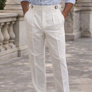 May include: White pleated trousers with a high waist and buckle details. The trousers are paired with a light blue button-down shirt and brown shoes. The image features a classic, tailored style.