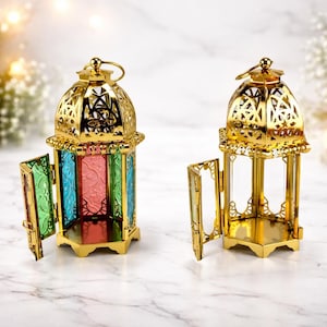 Peut inclure: Deux lanternes dorées ornées avec des portes ouvertes. Les lanternes ont des panneaux de vitrail colorés en vert, bleu et rouge. Le design comprend un dessus décoratif avec une boucle pour suspendre. Les lanternes sont sur une surface en marbre.