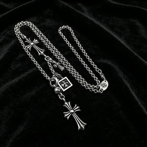 May include: A silver-toned necklace with multiple cross pendants and a square pendant. The necklace features a chain design and is displayed on a black surface. The crosses have detailed designs.