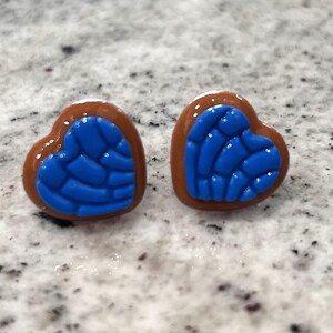 Op de afbeelding: Een paar hartvormige oorstekers. Elke oorbel heeft een blauw, gestructureerd ontwerp binnen een bruine rand. De oorbellen zijn geplaatst op een gespikkeld, lichtgekleurd oppervlak.