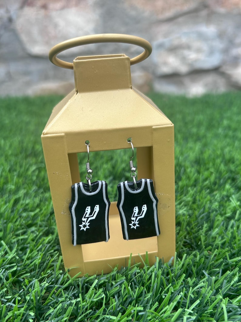 May include: A pair of black and white basketball jersey earrings. The earrings are shaped like miniature jerseys with the Spurs logo and silver hooks. The earrings are displayed inside a gold lantern, set against green grass.