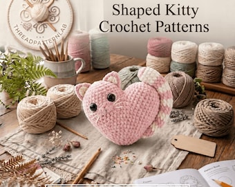 Puede incluir: Un peluche de gato tejido a ganchillo en forma de corazón rosa con grandes ojos negros y bigotes blancos, rodeado de ovillos de lana, ganchillos de madera, flores secas y un libro de patrones con el texto Celia the Heart-Shaped Kitty Crochet Patterns.