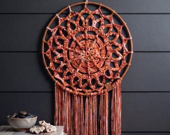 Könnte beinhalten: Ein kreisförmiger Wandbehang mit einem terrakottafarbenen Häkel-Mandala in einem Holzring mit langen, herabhängenden Garnfransen. Der Text lautet: Terracotta Sunset Mandala Wall Hanging Crochet Pattern. Instant PDF Download.