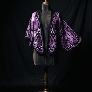 Puede incluir: Un chal de ganchillo dise&ntilde;ado para parecerse a alas de mariposa en tonos morado, lavanda y negro. La prenda presenta patrones de punto intrincados, bordes festoneados y peque&ntilde;os acentos circulares blancos a lo largo de los paneles.