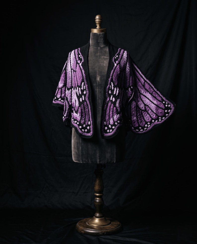 Puede incluir: Un chal de ganchillo dise&ntilde;ado para parecerse a alas de mariposa en tonos morado, lavanda y negro. La prenda presenta patrones de punto intrincados, bordes festoneados y peque&ntilde;os acentos circulares blancos a lo largo de los paneles.
