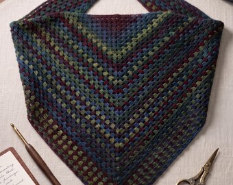 Könnte beinhalten: Ein dreieckiges Häkeltuch mit einem variierenden Muster aus dunkelburgunderfarbenem, waldgrünem und marineblauem Garn. Der Text lautet Midnight Woods Shawl Crochet Pattern und Instant PDF Download. Eine Häkelnadel aus Holz und eine Metallschere liegen bei.