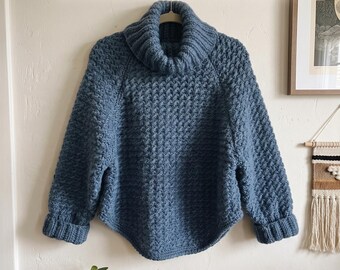May include: A blue crochet sweater dress with a thick turtleneck collar, long sleeves with ribbed cuffs, and a textured stitch pattern. The image includes the text: Crochet Cloud Wrap Sweater Dress Pattern and Instant PDF Download.