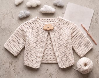 May include: A cream-colored crochet cardigan with a single cloud-shaped wooden button closure. The text reads The Little Cloud Cardigan Crochet Pattern and Instant PDF Download. Includes a ball of cream yarn, a crochet hook, and small felt clouds.