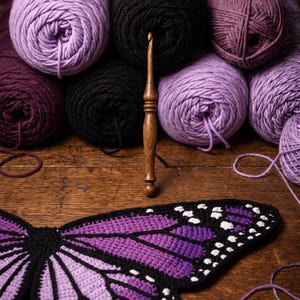 Puede incluir: Un ganchillo de madera se encuentra en posici&oacute;n vertical frente a varios ovillos de lana en tonos morado, negro y ciruela. Debajo de la lana, un ala de mariposa tejida a ganchillo en degradado morado, negro y blanco sobre madera.