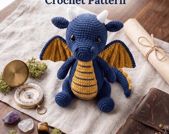Könnte beinhalten: Ein dunkelblaues, gehäkeltes Amigurumi-Drachenspielzeug mit weißen Hörnern, gelben Flügeln und einem gelben Bauch. Der Text lautet: Cute Blue Dragon Amigurumi Toy Crochet Pattern. Instant PDF Download. Der Drache ist von einem Kompass, Steinen und einer Schriftrolle umgeben.