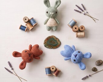 Könnte beinhalten: Drei gehäkelte Hasen in Salbeigrün, Rostorange und Hellblau, begleitet von hölzernen Garnrollen, getrockneten Lavendelzweigen und einer polierten Achat-Scheibe. Der Text lautet Crochet Rustic Stitch Bunnies Pattern und Instant PDF Download.