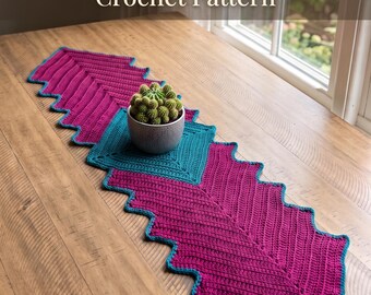 Könnte beinhalten: Ein gehäkelter Tischläufer mit Chevron-Pfeilmuster in Magenta und Türkis. Der Text "Chevron Arrow Table Mat Crochet Pattern" und "Instant PDF Download" ist sichtbar. Ein kleiner grüner Kaktus in einem grauen Topf steht auf dem Läufer.