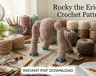 May include: A crocheted creature with a textured dusty pink body and small teal claws, surrounded by various yarn balls in neutral tones, wooden crochet hooks, dried flowers, and an open instruction booklet labeled Rocky the Eridian Crochet Pattern.