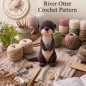 Puede incluir: Una nutria de río de ganchillo marrón oscuro y beige que sostiene una pequeña piedra gris, rodeada de ovillos de lana, ganchillos de madera, flores secas y un libro con el texto The Little River Otter Crochet Pattern e INSTANT PDF DOWNLOAD.